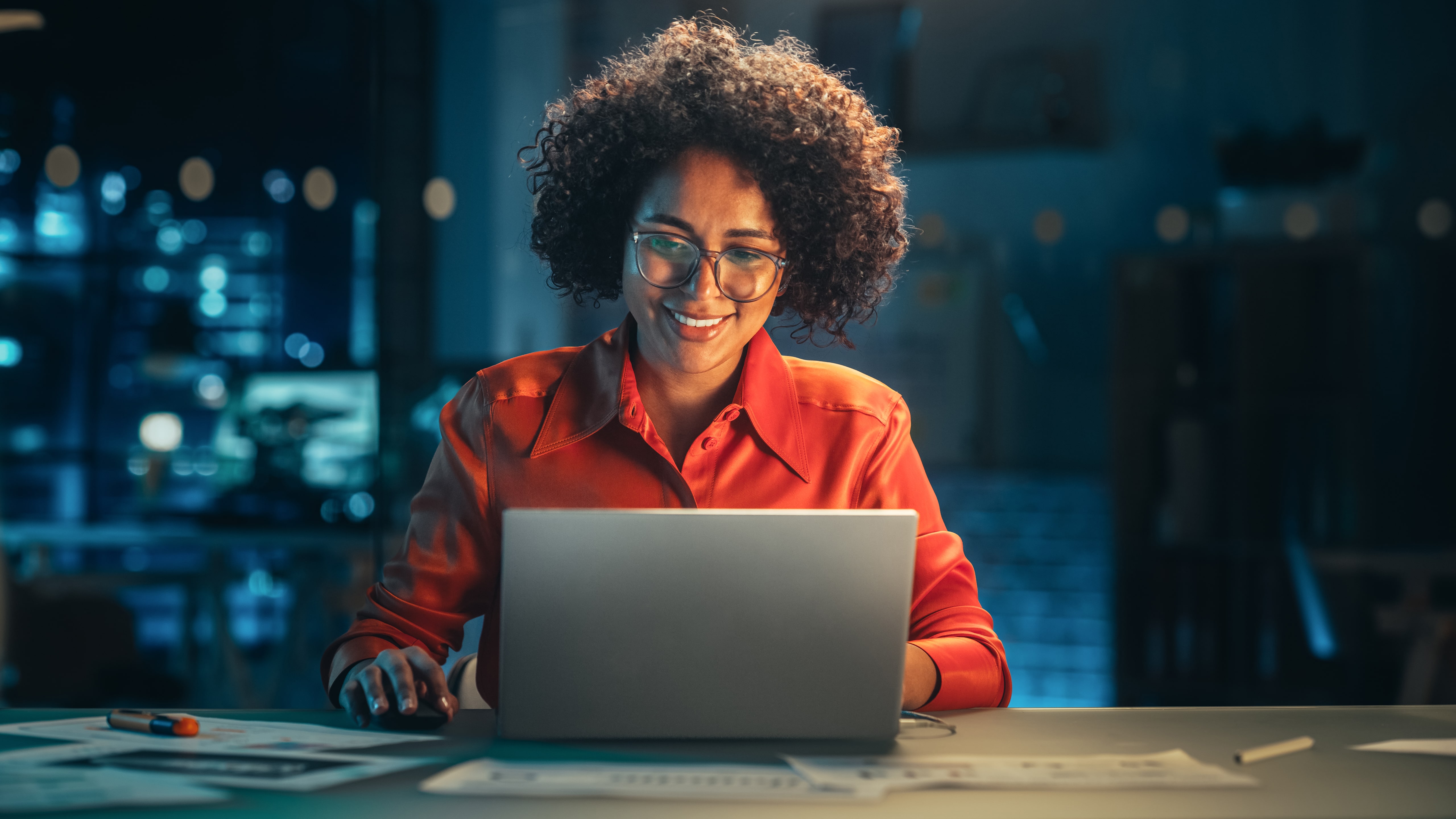 Woman in orange blouse working at laptop at night, with city light in background through window